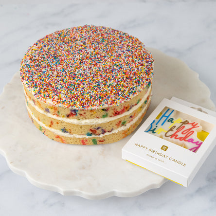 three-tiered Sprinkle layer cake topped with rainbow sprinkles with a happy birthday block candle laying on the side